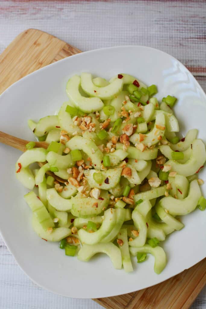 A white plate with a salad of sliced celery and chopped nuts, reminiscent of a thai cucumber salad and garnished with red pepper flakes, rests on a wooden cutting board. A wooden fork is on the plate.