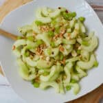 A white bowl filled with Thai cucumber salad—sliced celery, cucumber, and chopped nuts—rests on a wooden board with a wooden spoon on the side, all set atop a white surface.