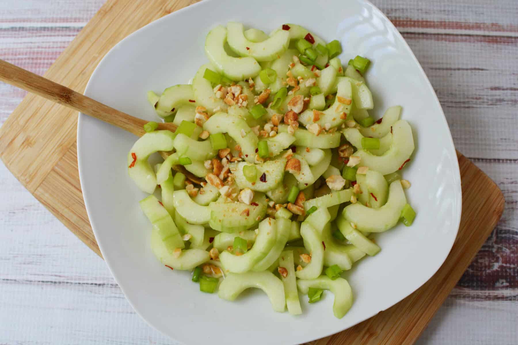 A white bowl filled with Thai cucumber salad—sliced celery, cucumber, and chopped nuts—rests on a wooden board with a wooden spoon on the side, all set atop a white surface.