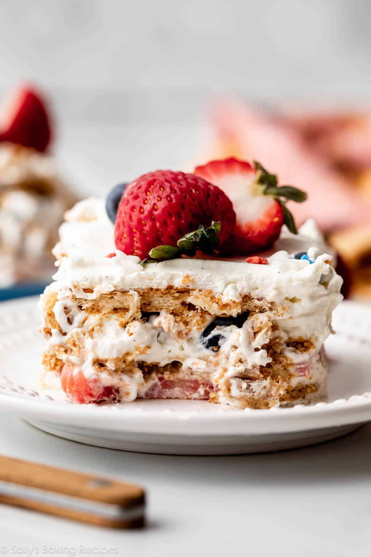 A slice of layered dessert with whipped cream, berries, and graham crackers sits on a white plate, topped with a whole strawberry and blueberry.