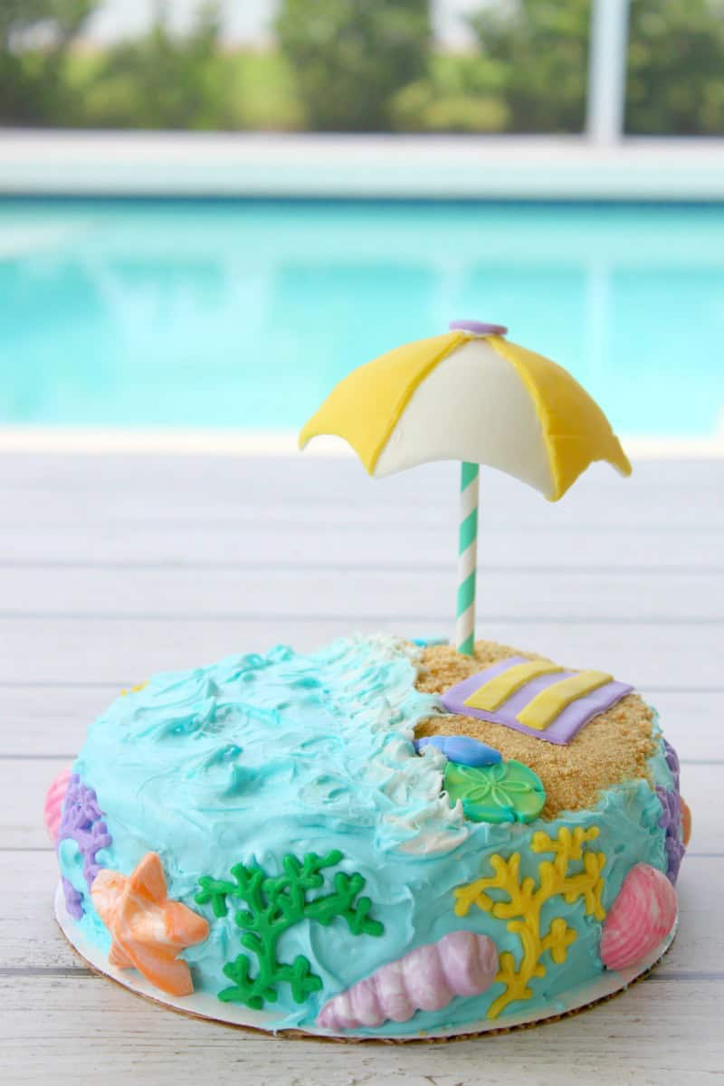A round cake decorated like a beach scene, with blue icing for water, seashells, starfish, and coral—a perfect choice for summer birthday cakes—features a small towel, sandy area, and a yellow-and-white umbrella beside a swimming pool.