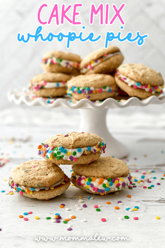 A plate of colorful Cake Mix Whoopie Pies filled with frosting and rolled in rainbow sprinkles sits on a cake stand, with extra Cake Mix Whoopie Pies and scattered sprinkles on the white surface below.