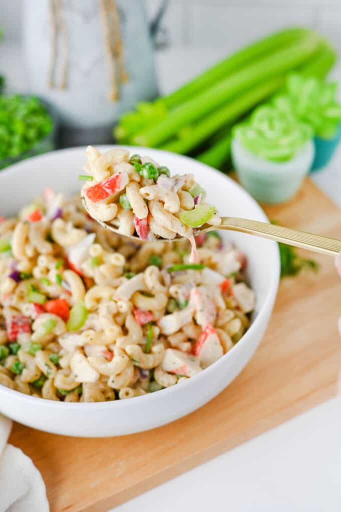 A close-up of a fork lifting a bite of creamy Crab Pasta Salad with vegetables from a white bowl, with fresh celery and greens blurred in the background.