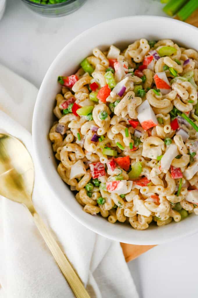A white bowl filled with creamy Crab Pasta Salad featuring chopped red peppers, celery, red onion, and green herbs sits on a white surface next to a gold spoon and a beige napkin.