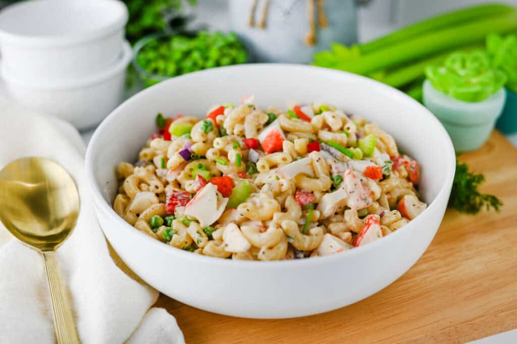A white bowl filled with Crab Pasta Salad—featuring pasta, diced crab sticks, celery, green onions, and peas—rests on a wooden surface beside a gold spoon and white napkin. Fresh vegetables appear in the background.