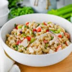 A white bowl filled with Crab Pasta Salad—featuring pasta, diced crab sticks, celery, green onions, and peas—rests on a wooden surface beside a gold spoon and white napkin. Fresh vegetables appear in the background.