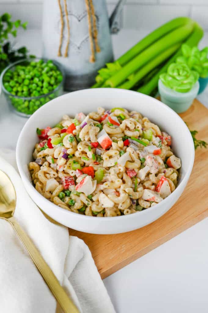 A white bowl filled with creamy Crab Pasta Salad featuring chopped celery, red bell pepper, peas, and imitation crab. Fresh celery stalks, peas, and a gold spoon sit nearby on a white countertop.