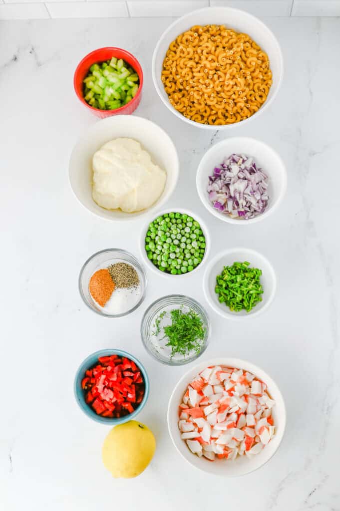 An overhead view of bowls with ingredients for a Crab Pasta Salad, including elbow macaroni, celery, mayonnaise, red onion, peas, green onions, spices, diced red pepper, dill, imitation crab, and a lemon on a white surface.