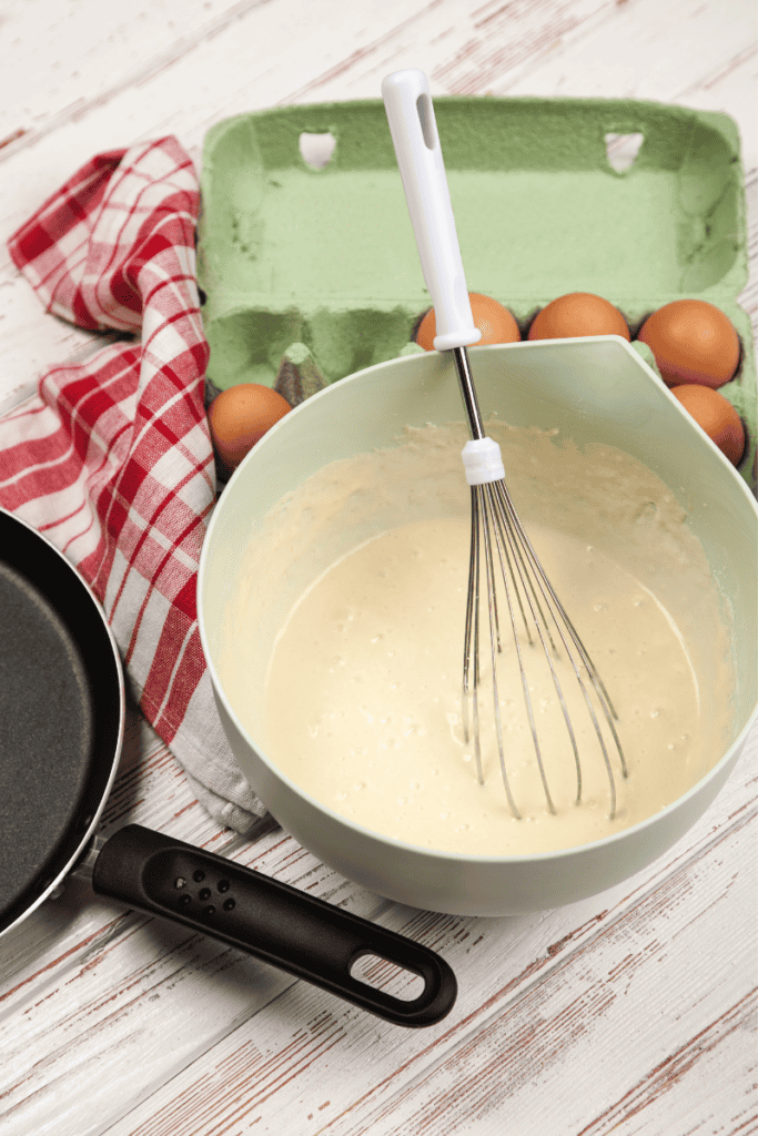 A mixing bowl with batter and a whisk, eggs in a carton, a frying pan, and a red and white checked towel on a wooden surface—perfect inspiration when wondering what to do with leftover pancake batter.