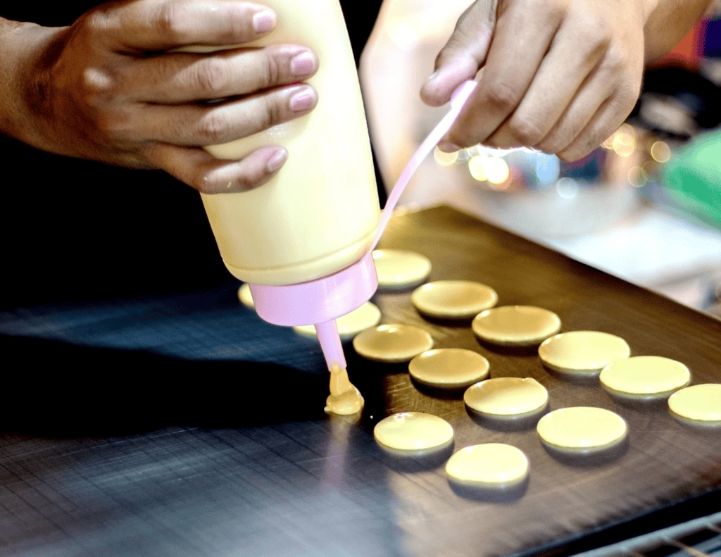 A person squeezes creamy batter from a bottle onto a baking tray, forming small round shapes for cookies or pastries—a perfect idea for what to do with leftover pancake batter.