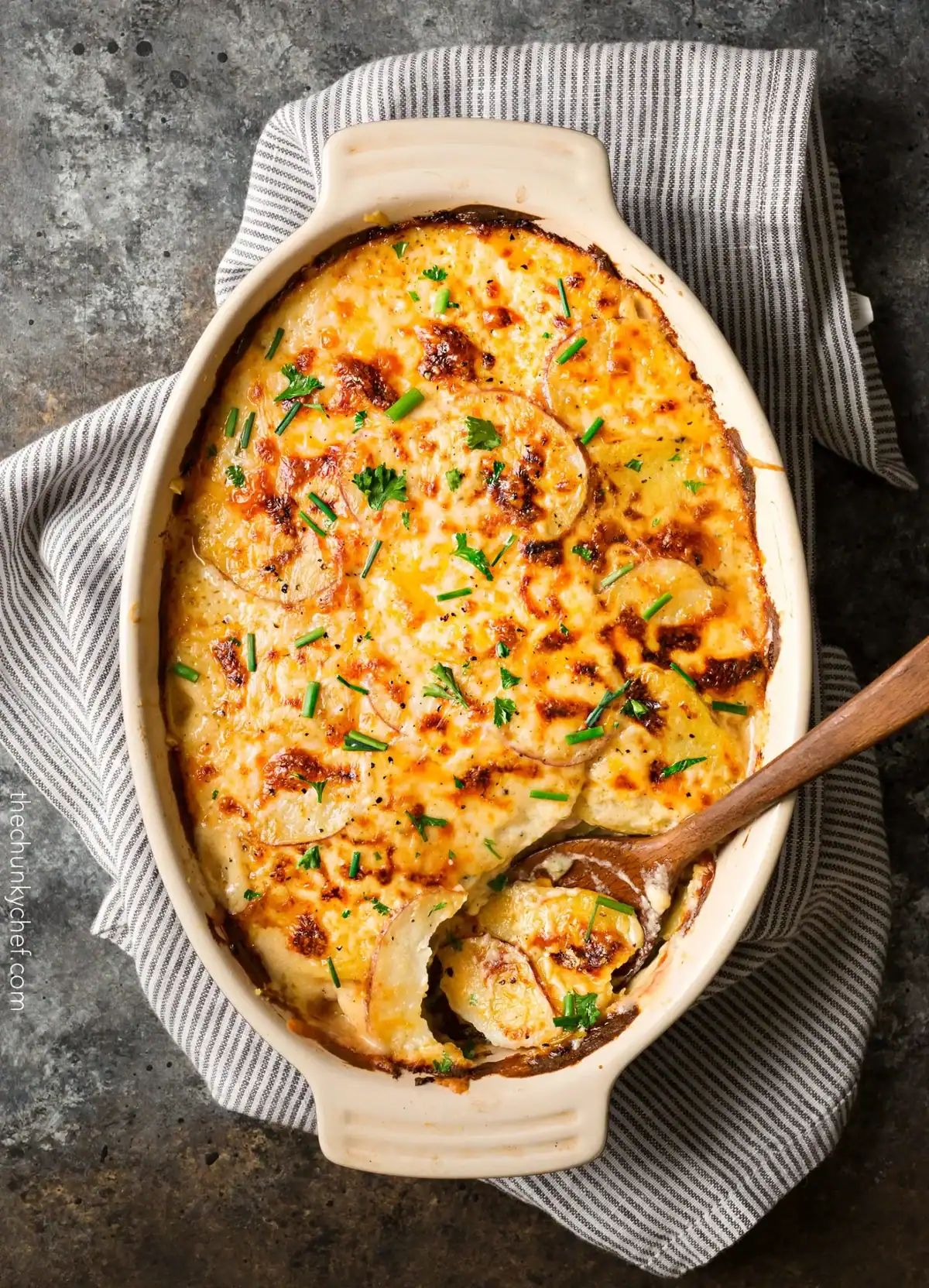 A baked dish of creamy potato gratin topped with melted cheese and chopped chives, with a serving spoon and one portion removed. The dish rests on a striped cloth on a dark stone surface.