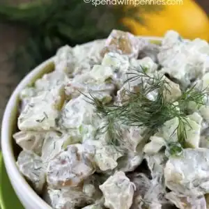 A bowl of creamy potato salad garnished with fresh dill, with a sprig of dill and a lemon in the background.