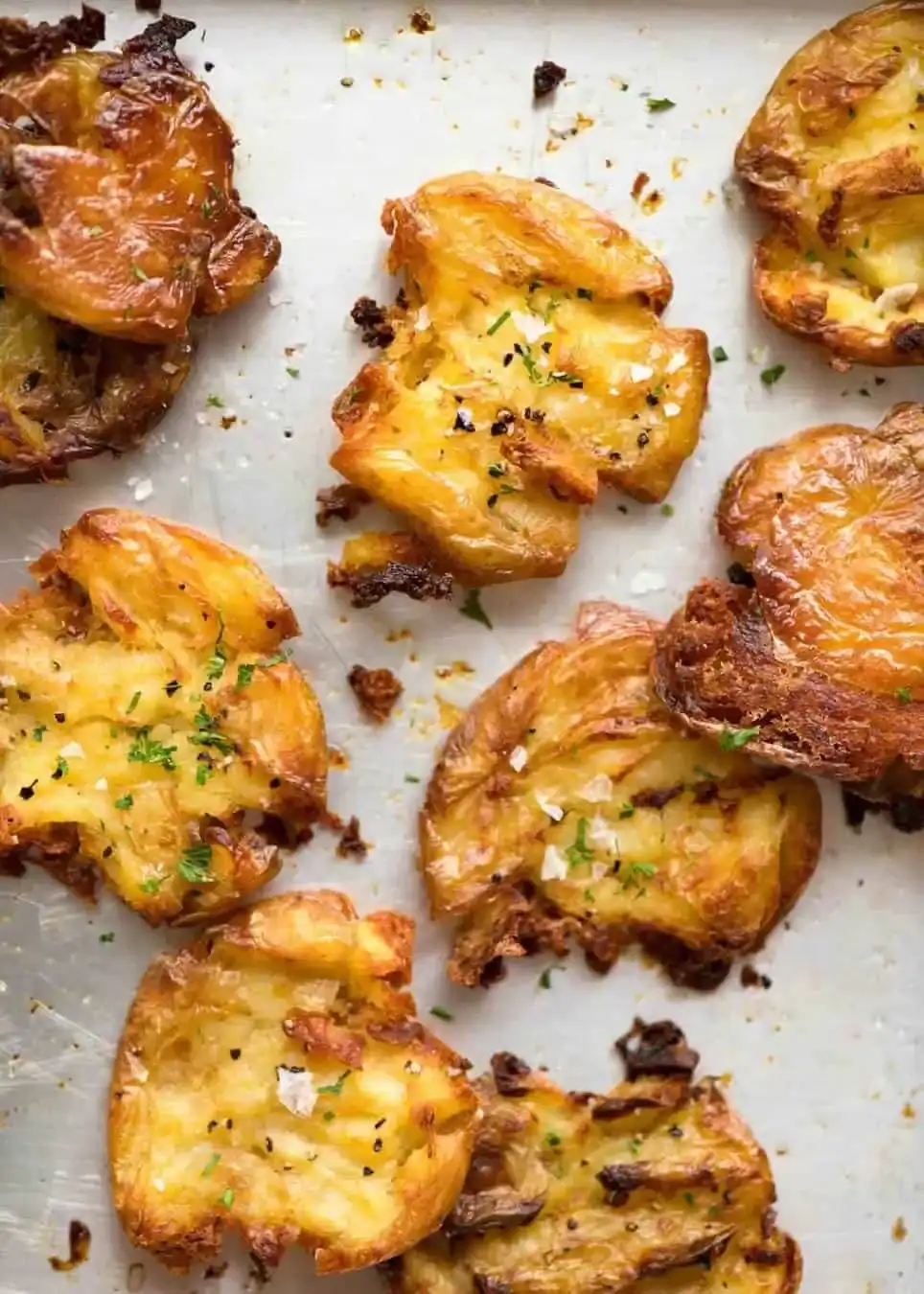 Golden, crispy smashed potatoes topped with herbs and coarse salt, arranged on a metal baking sheet.