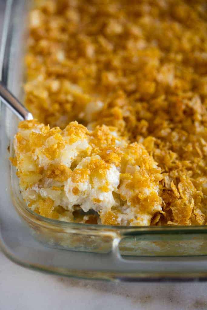 A close-up of a glass baking dish filled with cheesy hashbrown casserole topped with a crispy, golden cornflake crust. A serving spoon is scooping out a portion.