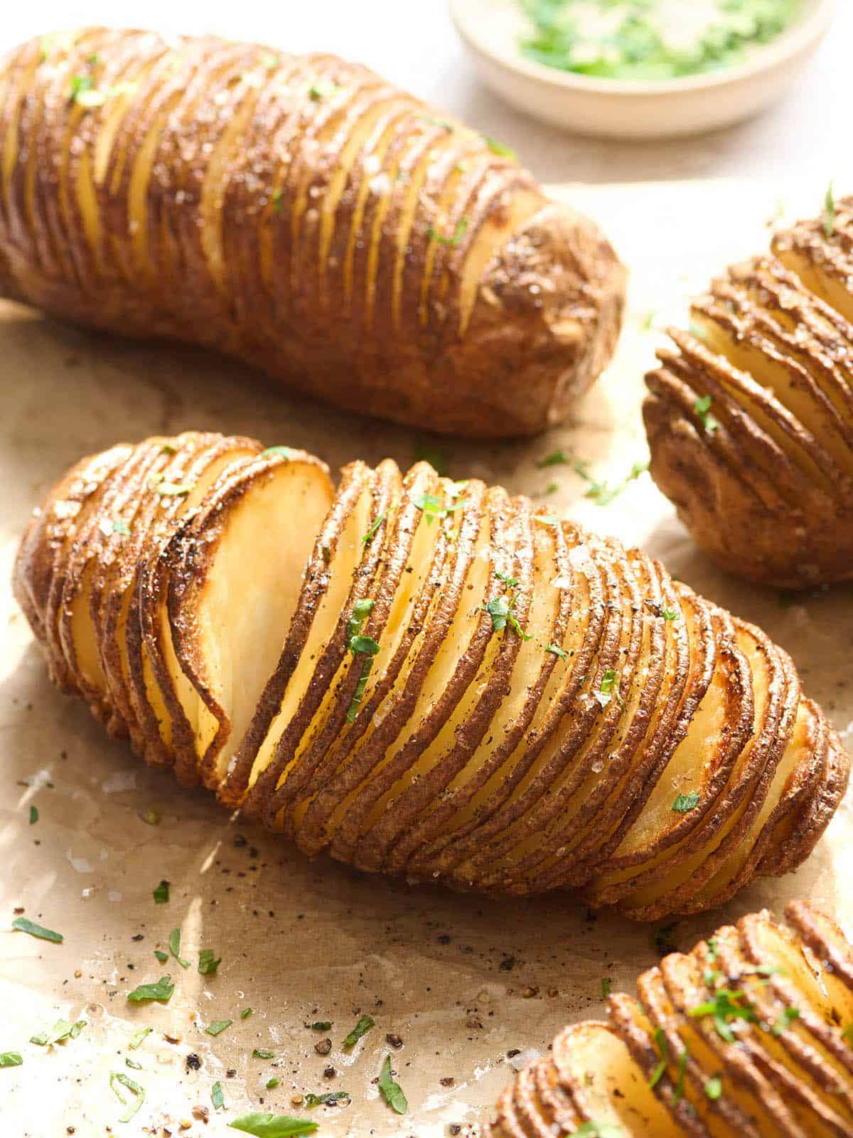 Three golden-brown Hasselback potatoes with thin, evenly sliced sections, sprinkled with chopped herbs and black pepper, rest on parchment paper. A small bowl of green garnish is visible in the background.