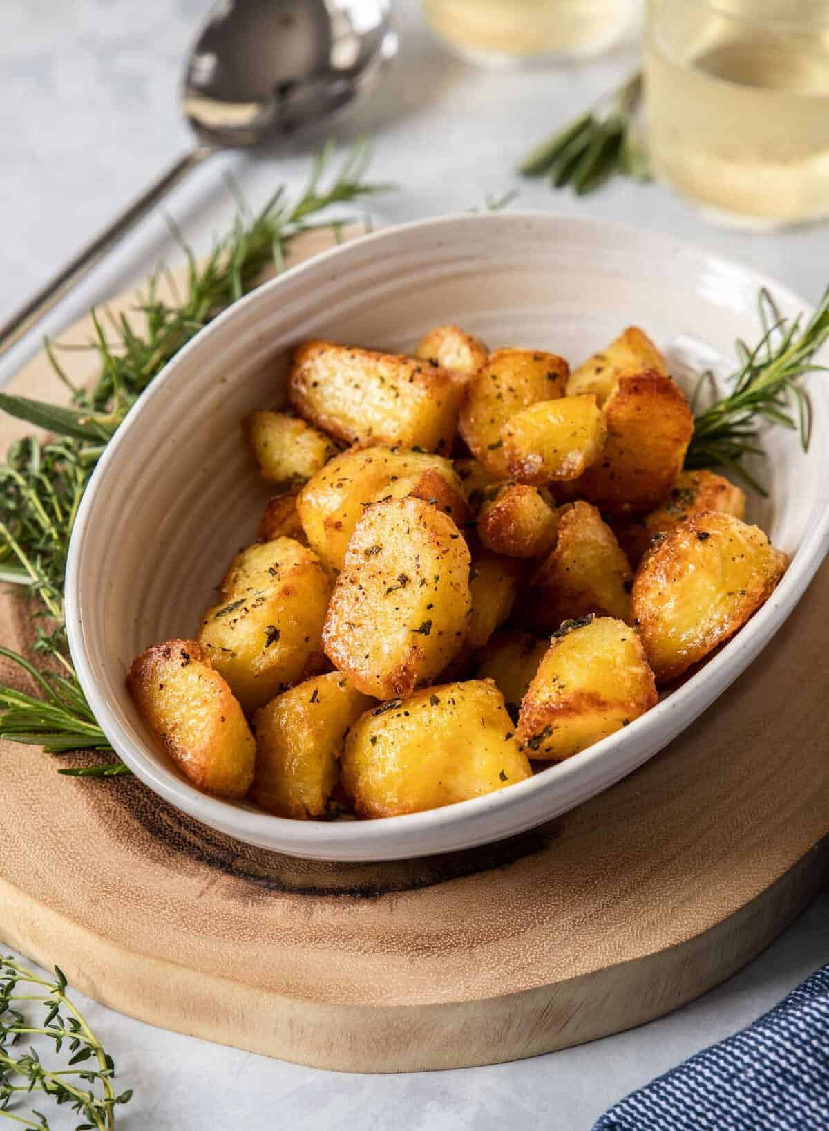 A white oval dish filled with golden, crispy roasted potatoes garnished with herbs sits on a wooden board, with sprigs of rosemary nearby and cutlery in the background.