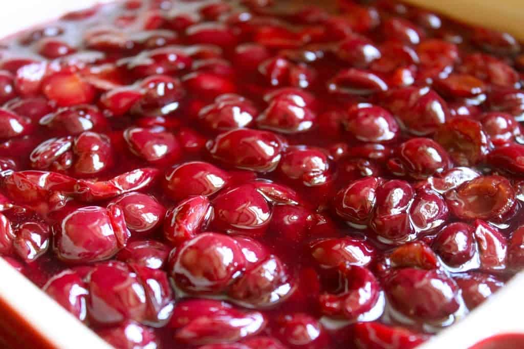 Close-up of glossy, cooked cherries in a thick, red sauce, filling a baking dish. The cherries look soft and juicy, coated in a shiny syrup—perfect inspiration for pie filling recipes or other delightful desserts.