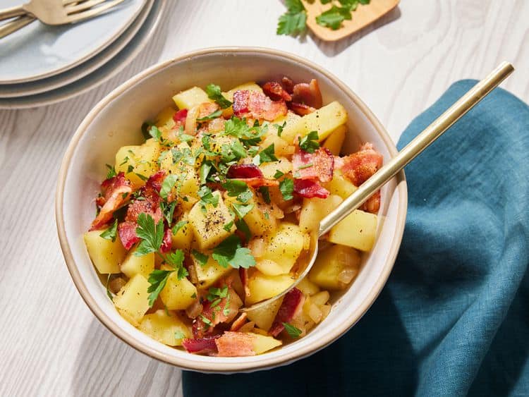 A bowl of potato salad topped with chopped bacon and fresh parsley, with a gold spoon resting inside. The bowl sits on a light wooden table next to a blue napkin and some stacked plates.