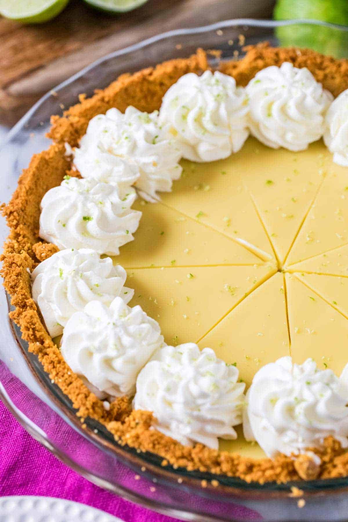 A key lime pie with a graham cracker crust, inspired by classic Pie Filling Recipes, is topped with whipped cream rosettes and lime zest as it sits on a table. The pie is pre-sliced, and a magenta cloth is partially visible underneath the pie dish.