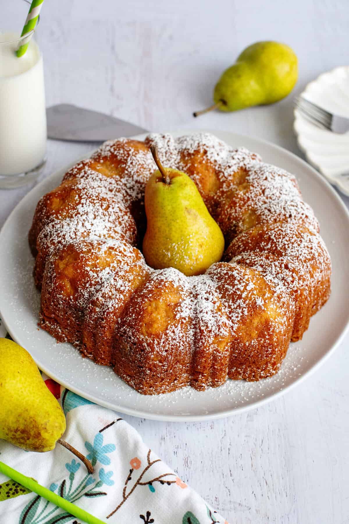 A classic example of fall bundt cake recipes, this bundt cake dusted with powdered sugar sits on a white plate, topped with a whole pear in the center. Fresh pears and a glass of milk are nearby on a white table.