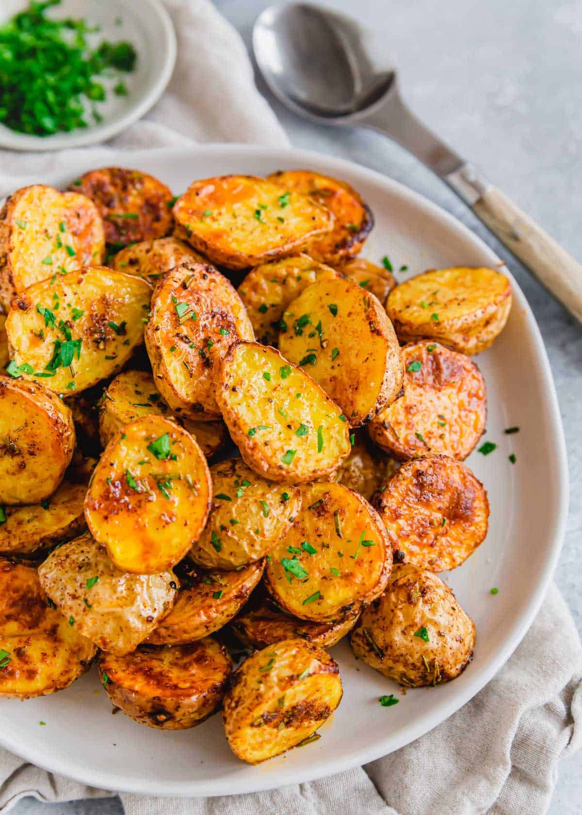 A plate of golden, roasted baby potatoes garnished with chopped herbs, with a spoon and a small bowl of green herbs in the background.