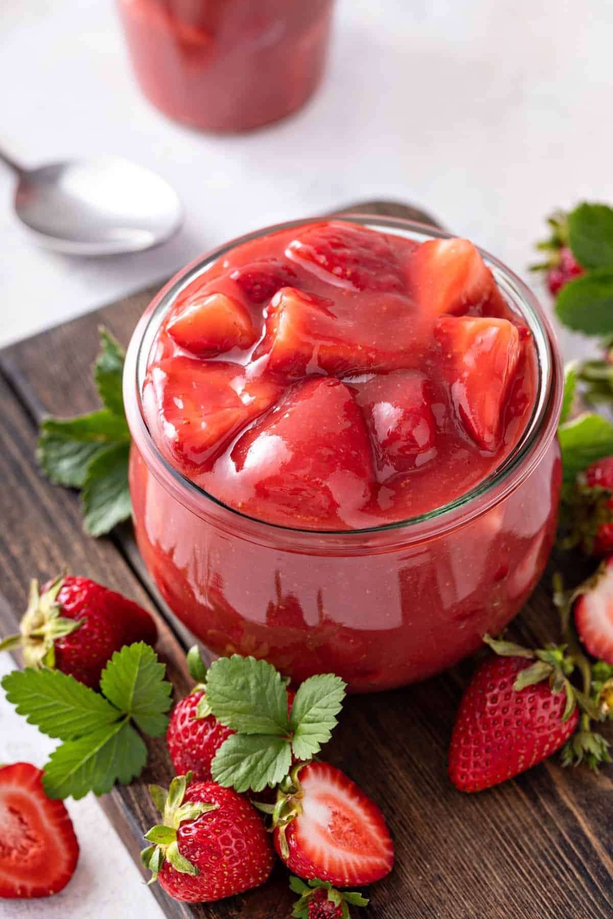 A glass jar filled with chunky strawberry compote sits on a wooden board, surrounded by fresh strawberries and green leaves, offering inspiration for pie filling recipes. A spoon and another jar are blurred in the background, highlighting the homemade charm of this delicious scene.