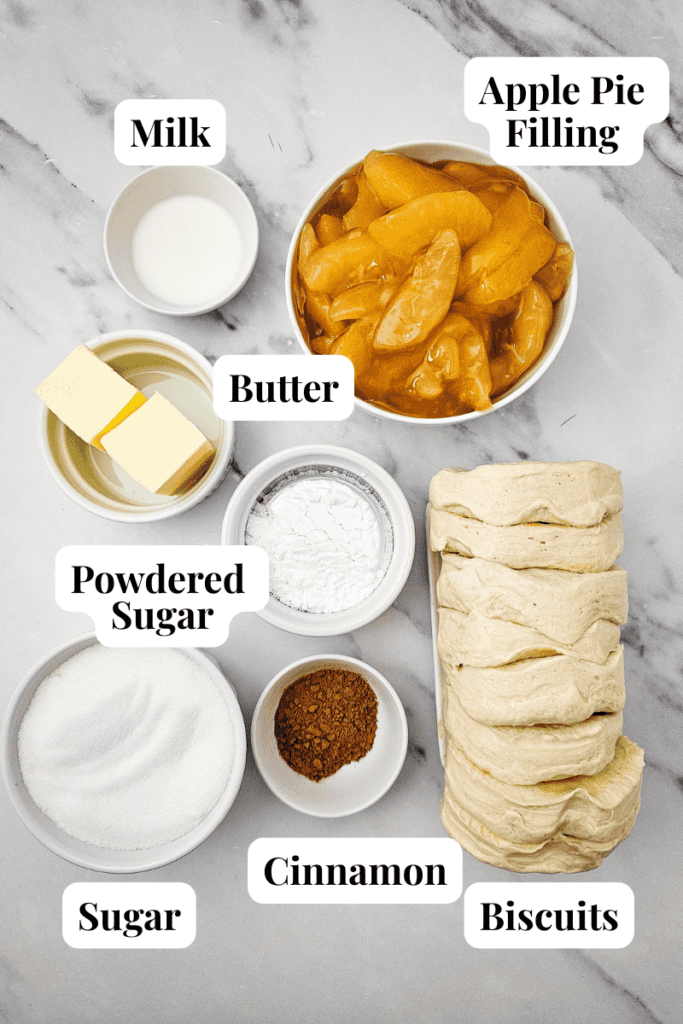 Bowls of milk, apple pie filling, butter, powdered sugar, sugar, cinnamon, and a stack of biscuit dough are arranged on a marble surface—each labeled with its name—ready to be transformed into warm and delicious Apple Cinnamon Pull Apart Bread.