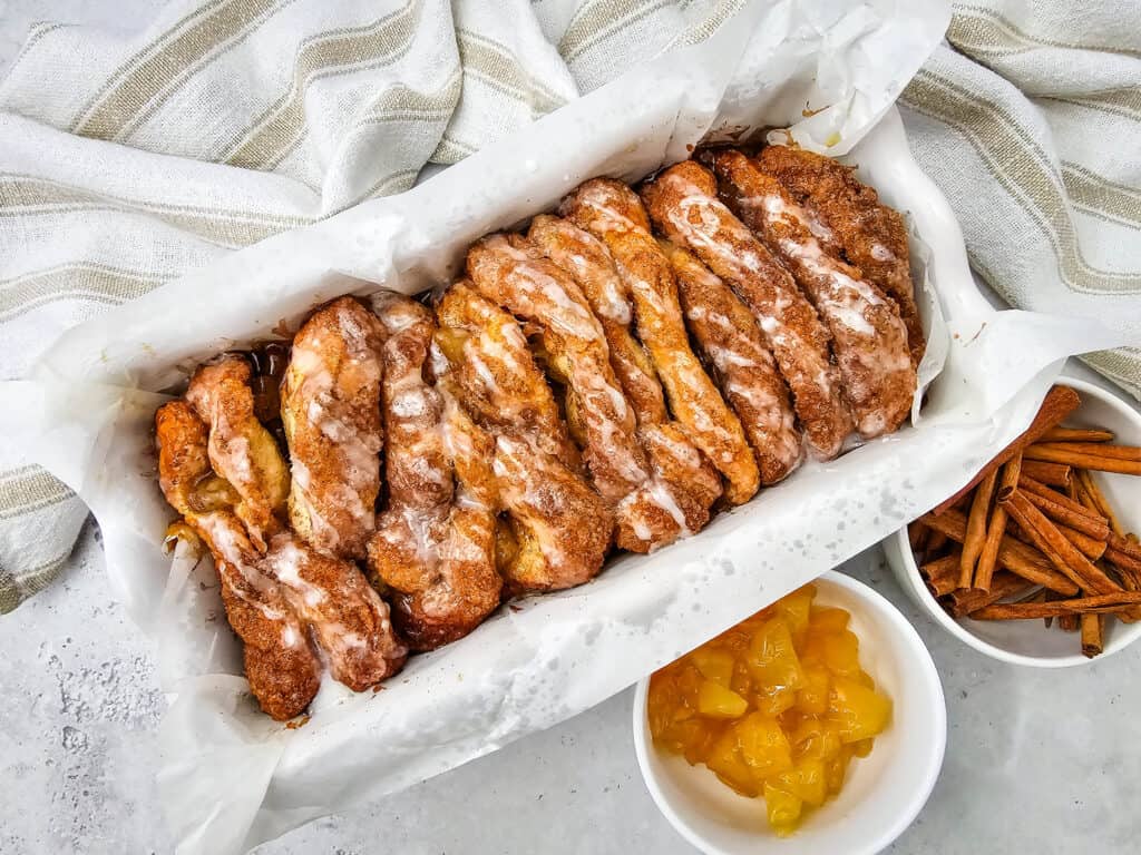 A loaf of Apple Cinnamon Pull Apart Bread, glazed to perfection in a parchment-lined pan, sits beside bowls of diced pineapple and cinnamon sticks on a light surface with a striped towel.