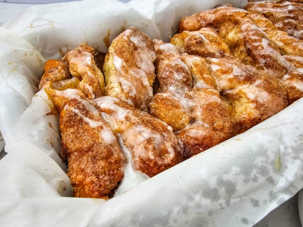 Freshly baked Apple Cinnamon Pull Apart Bread with a sugar glaze sits in a parchment-lined pan, featuring golden brown, twisted sections and a generous coating of cinnamon and icing.