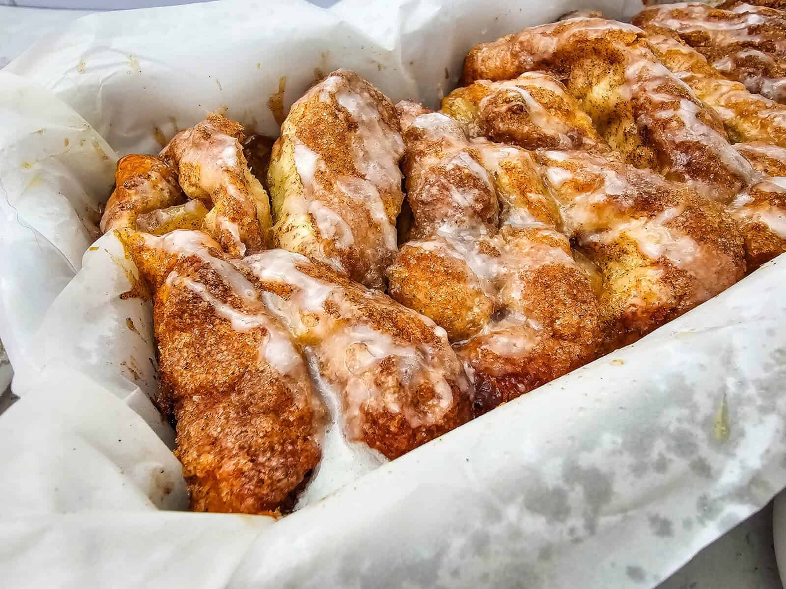 Freshly baked Apple Cinnamon Pull Apart Bread with a sugar glaze sits in a parchment-lined pan, featuring golden brown, twisted sections and a generous coating of cinnamon and icing.