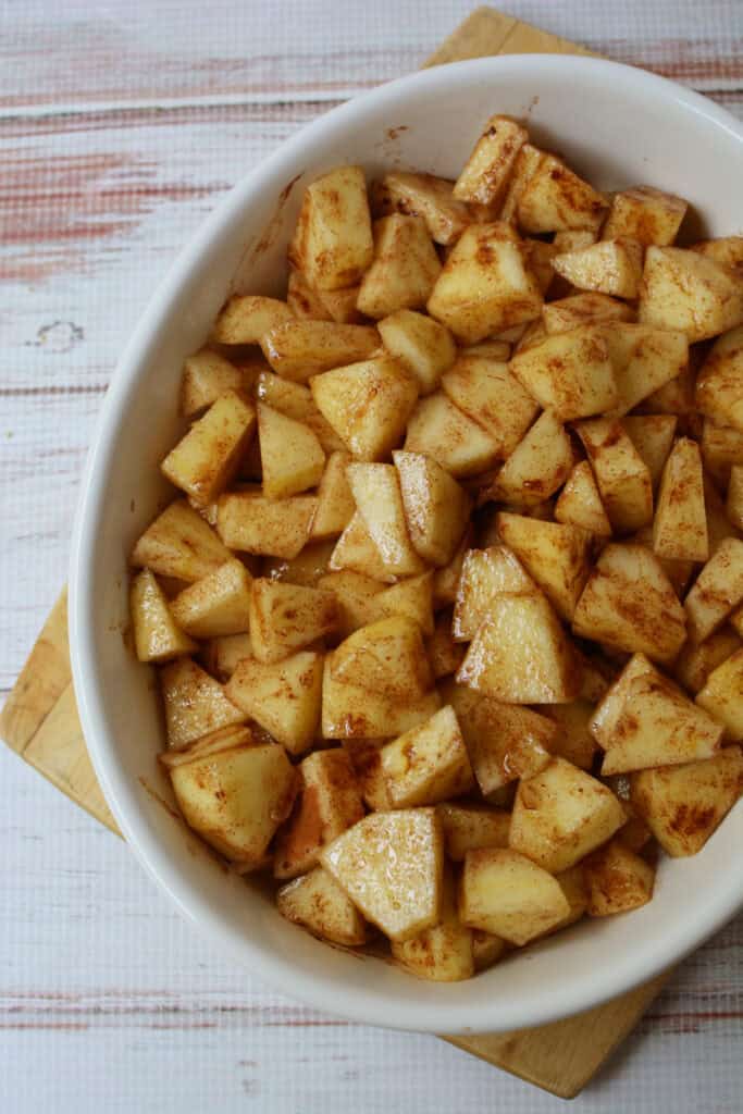 A white oval baking dish filled with chopped apples coated in cinnamon and sugar sits on a light wooden surface, ready to be transformed into delicious Baked Cinnamon Apples.