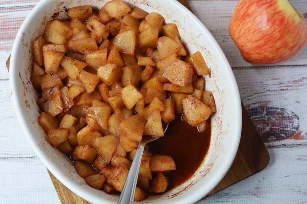 A white dish filled with Baked Cinnamon Apples, diced and coated in syrup, sits on a wooden board next to a fresh red apple. A metal spoon rests inside the dish.