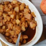 A white dish filled with Baked Cinnamon Apples, diced and coated in syrup, sits on a wooden board next to a fresh red apple. A metal spoon rests inside the dish.