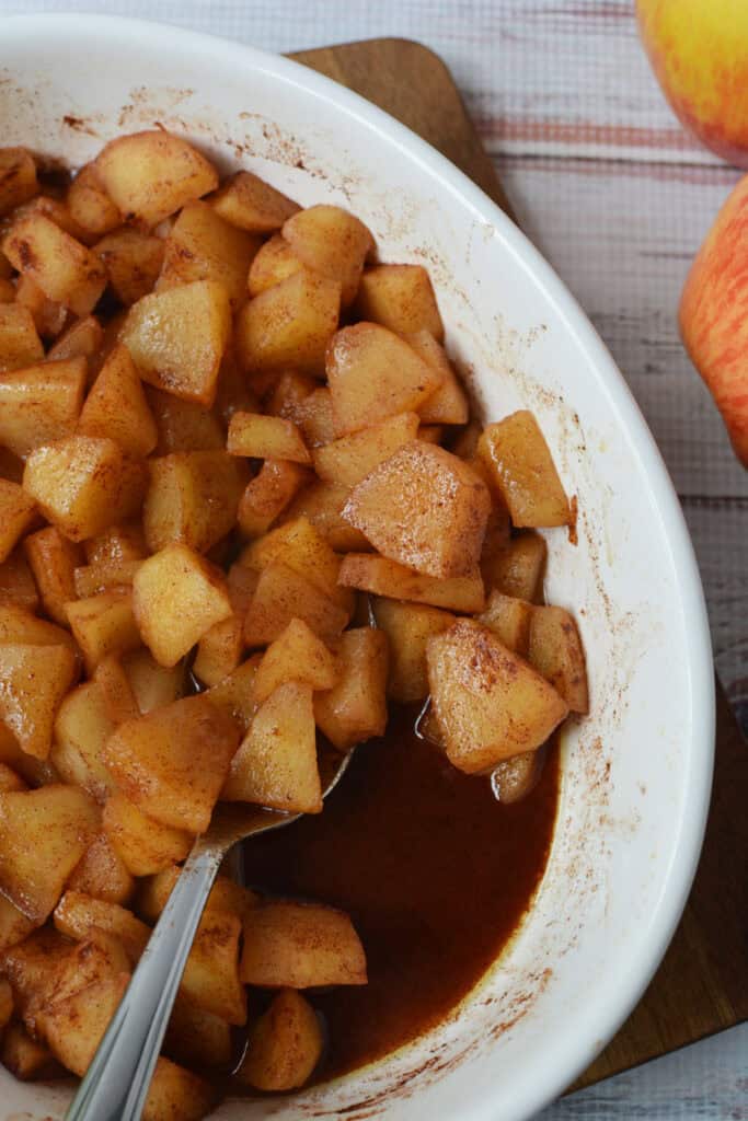 A white dish filled with Baked Cinnamon Apples—diced apples coated in cinnamon and syrup—rests invitingly, with a spoon inside. Two whole apples sit nearby on a wooden surface.