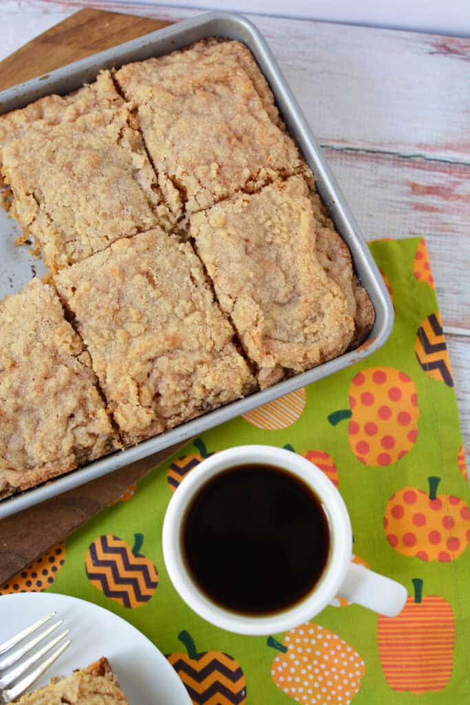A pan of crumb-topped Krusteaz Pumpkin Coffee Cake, cut into squares, sits beside a cup of black coffee on a napkin decorated with colorful, patterned pumpkins.