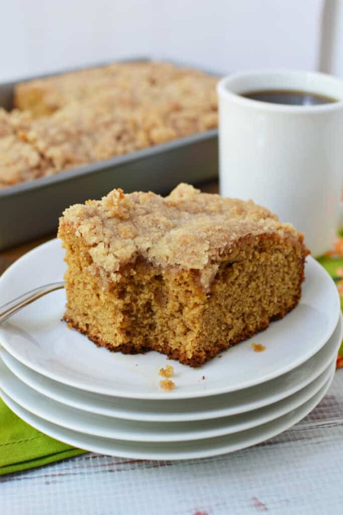 A slice of crumb-topped Krusteaz Pumpkin Coffee Cake rests on a stack of white plates, with a fork and a cup of black coffee beside it. In the background, a tray of similar cake awaits its turn to be enjoyed.