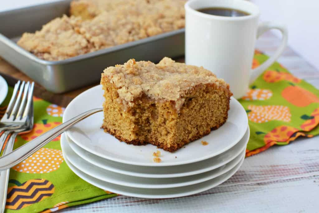 A slice of crumb-topped Krusteaz Pumpkin Coffee Cake sits on a stack of white plates with a fork, next to a mug of coffee and a pan of more cake, on a patterned green and orange napkin.