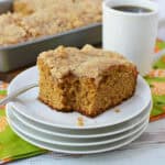 A slice of crumb-topped Krusteaz Pumpkin Coffee Cake sits on a stack of white plates with a fork, next to a mug of coffee and a pan of more cake, on a patterned green and orange napkin.