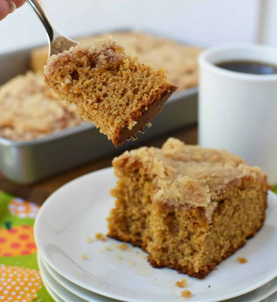 A hand holds a fork with a bite of crumb cake above a plate with a square piece of Krusteaz Pumpkin Coffee Cake; a mug of coffee and cake pan are in the background.