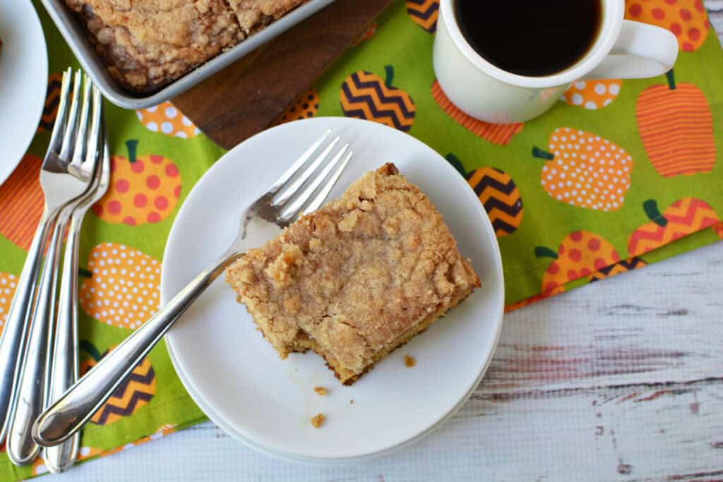 A plate with a slice of Krusteaz Pumpkin Coffee Cake and a fork sits next to a mug of black coffee. The tablecloth has orange pumpkins on a green background. There’s a pan with more cake in the corner.