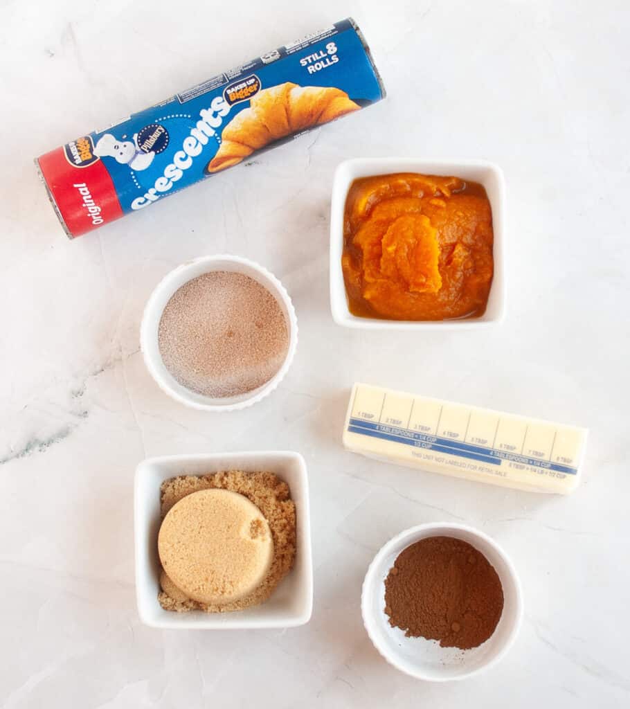 Ingredients on a white surface for Pumpkin Pie Twists: crescent roll dough, pumpkin puree, butter, brown sugar, cinnamon-sugar mix, and ground cinnamon, each in separate bowls or containers.