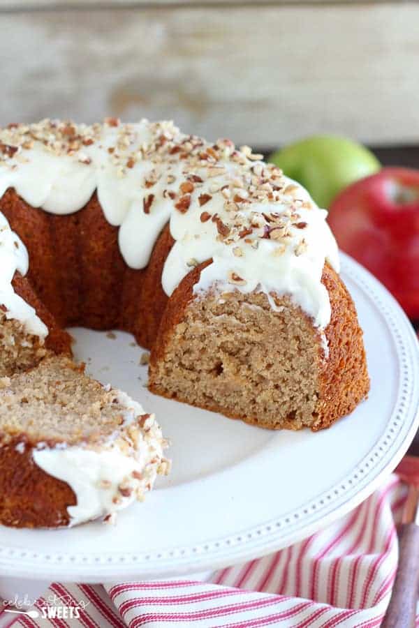 A spiced bundt cake with white icing and chopped nuts sits on a white cake stand. A slice has been removed. Apples and a green apple are visible in the background.
