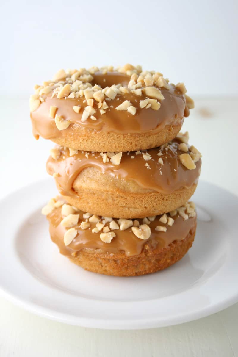 Three caramel-glazed donuts topped with chopped nuts are stacked on a white plate, set against a plain light background—perfect inspiration for your fall donut decorating ideas.