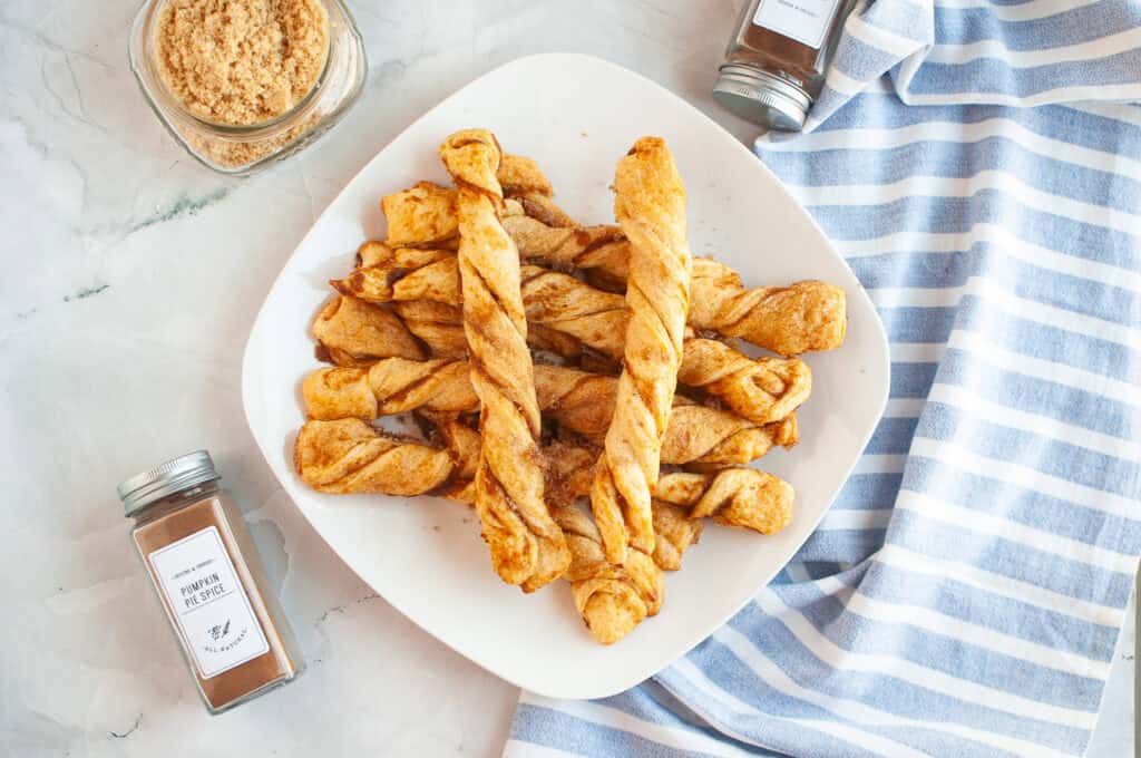 A plate of twisted Pumpkin Pie Twists sits on a marble surface near jars of pumpkin pie spice, brown sugar, and a blue striped towel.