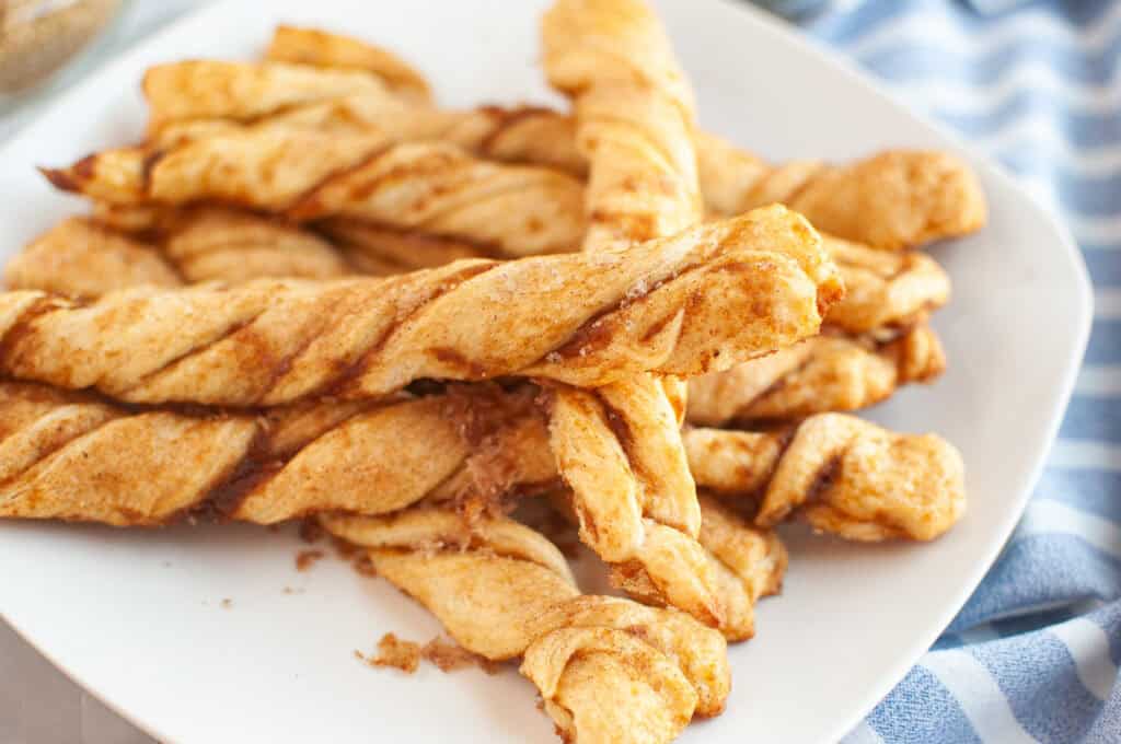 A white plate holds several golden brown Pumpkin Pie Twists with a cinnamon sugar coating, next to a blue and white striped cloth.