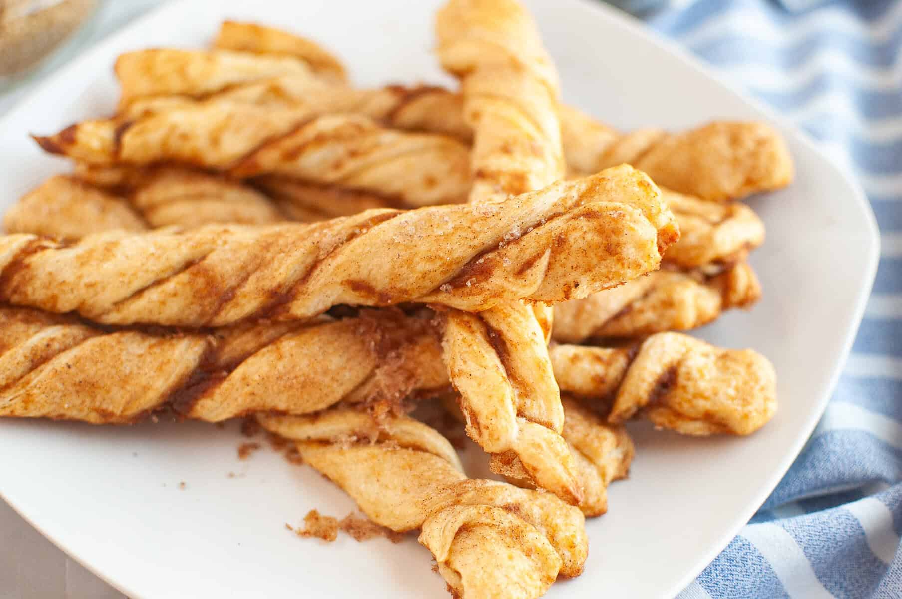 A white plate holds several golden brown Pumpkin Pie Twists with a cinnamon sugar coating, next to a blue and white striped cloth.