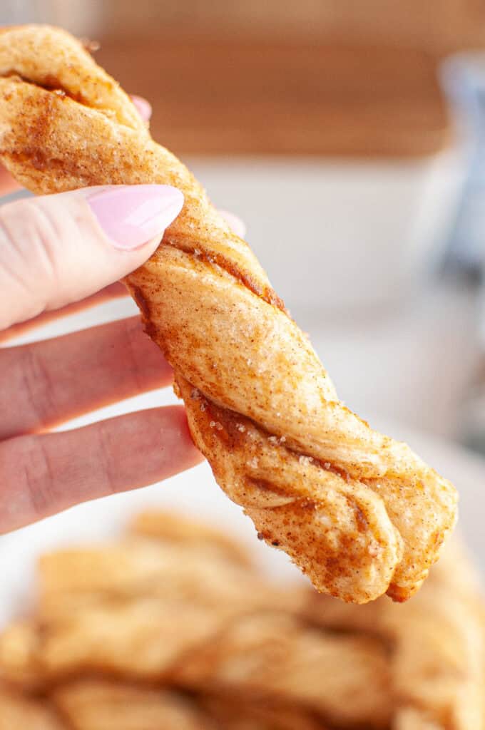 A hand holding a twisted Pumpkin Pie Twist coated with cinnamon and sugar, with more pastries blurred in the background.