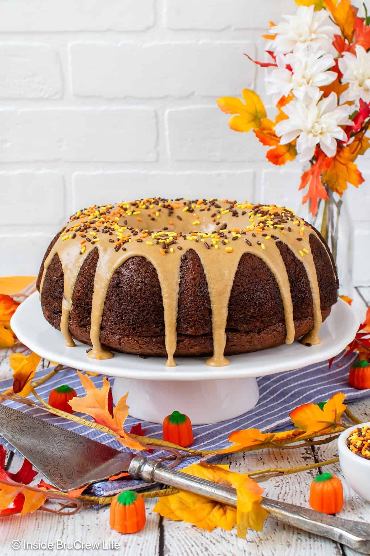 A chocolate bundt cake with tan glaze and fall-colored sprinkles on a white cake stand, surrounded by autumn leaves, candy pumpkins, and a bouquet of orange and white flowers.