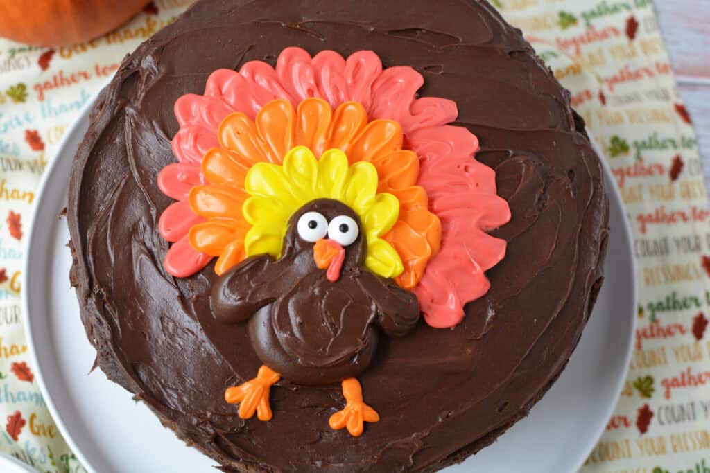 A Chocolate Thanksgiving Cake frosted with rich chocolate icing is decorated with a turkey made of colorful icing, featuring red, orange, and yellow feathers, and big eyes. The cake sits on a white plate against a festive Thanksgiving-themed background.