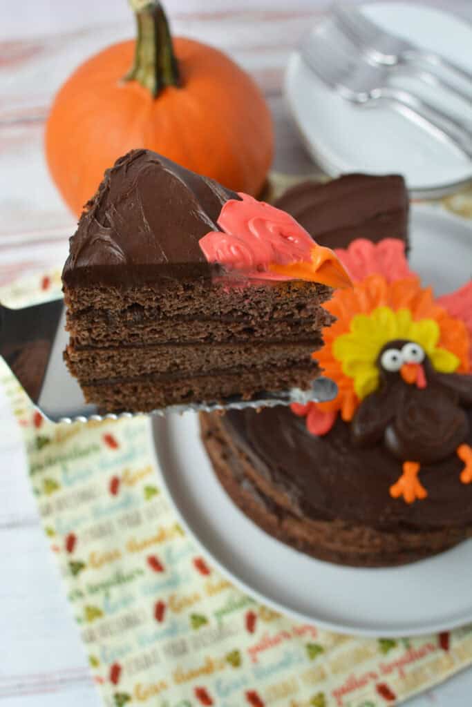 A slice of Chocolate Thanksgiving Cake with dark frosting is lifted from a decorated cake featuring a turkey made of icing. A small pumpkin and stacked forks are in the background.